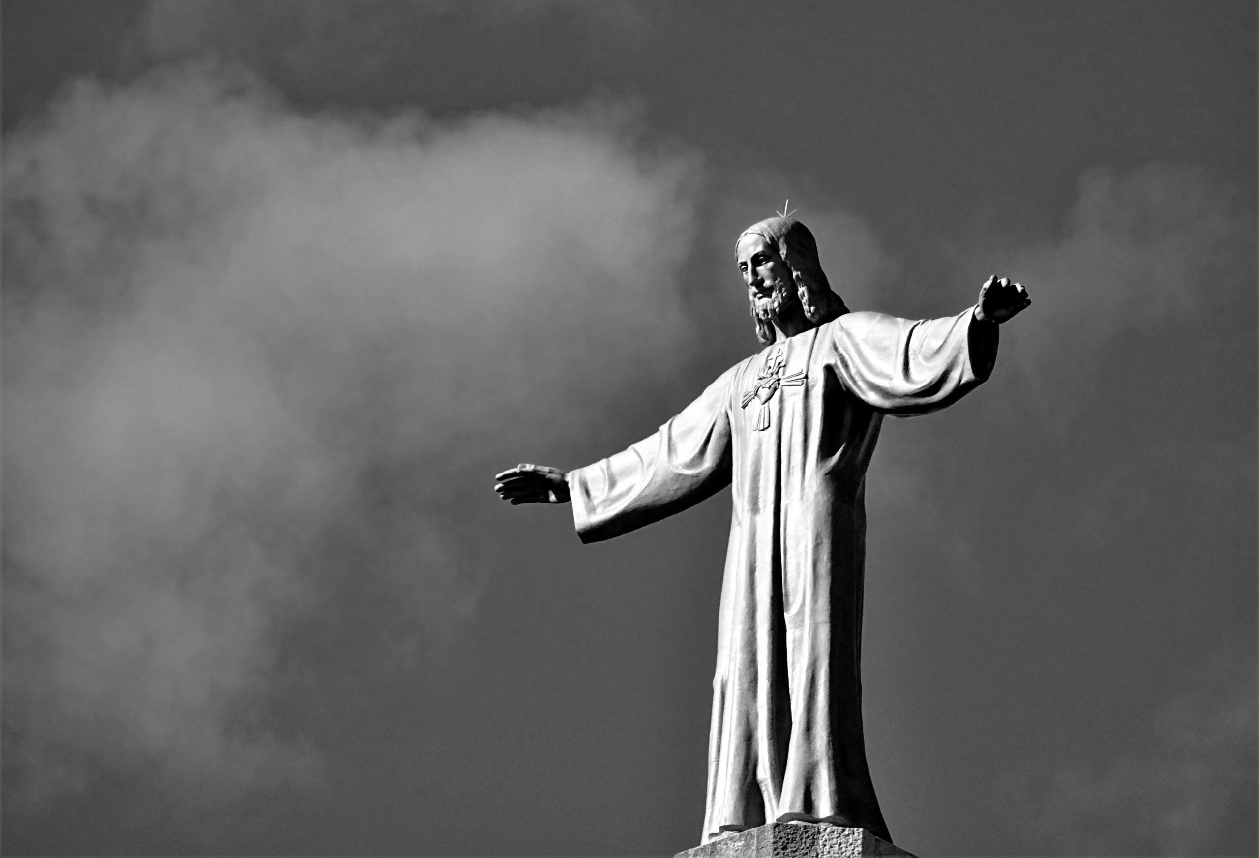 Black and white photo of a Jesus Christ statue reaching out against a cloudy sky.